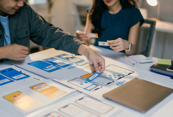 Two web designers are placing cutouts of buttons and icons onto a paper prototype of a mobile applic