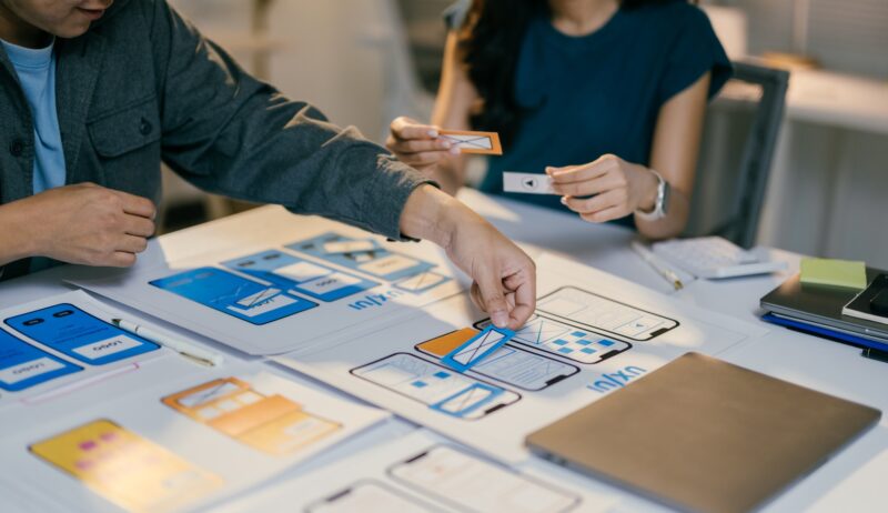 Two web designers are placing cutouts of buttons and icons onto a paper prototype of a mobile applic