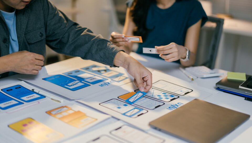 Two web designers are placing cutouts of buttons and icons onto a paper prototype of a mobile applic